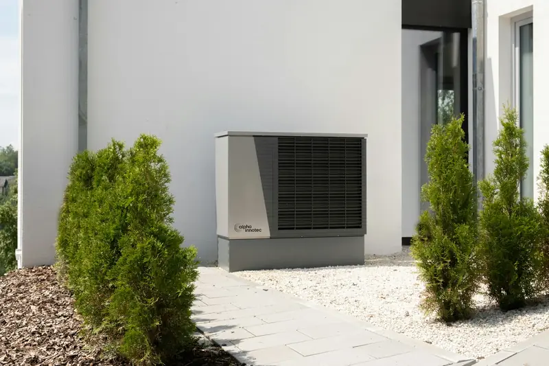 Outdoor heat pump unit beside a house wall between garden shrubs